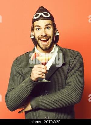 Schöner bärtiger Flieger mit langem Bart auf lächelndem Gesicht, der ein Glas alkoholischen Cocktail in grauem Strickpullover mit Hut und Brille auf rotem Studiohintergrund hält Stockfoto