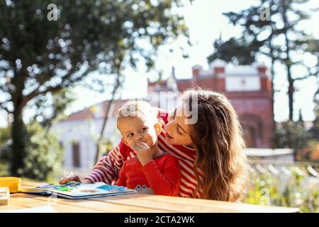 Glückliche Mutter und Kleinkind Lesen Lift-the-Flap Kinder Buch und Essen Snacks im Garten Stockfoto