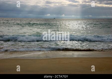 Dramatischer wolkig Sonnenuntergang mit Sonnenstrahlen über der Atlantikküste. Fischerschiff segelt am Horizont unter Sonnenstrahlen, die durch die Wolken leuchten. Nazare, Por Stockfoto