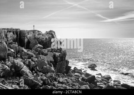 Überqueren Sie den felsigen Gipfel über den Ozean. Kreuz in den Himmel - Ebene Spur. Peniche, Portugal. Gealtertes Foto. Schwarz und Weiß. Stockfoto