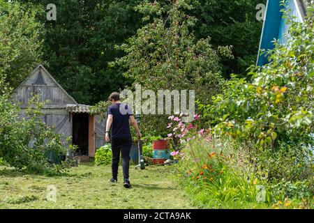 Ein junger Mann geht mit einem Trimer auf dem geschrubbelten Rasen. Stockfoto