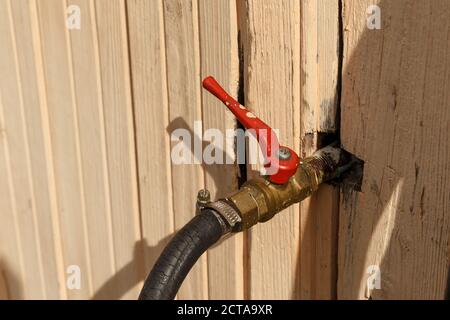 Der Wasserhahn ragt aus der Wand des Hauses. Ein Wasserhahn aus Eisen mit rotem Griff und schwarzem Schlauch. Stockfoto