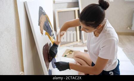 Konzentrierte Brünette mit Brötchen in weißem T-Shirt und schwarzen Shorts Zeichnet Bild laufende Hände mit Farben auf Leinwand in der Werkstatt Im Sommer Stockfoto