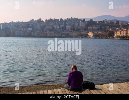 Der Mann entspannt sich auf dem hölzernen Pier mit Panoramablick auf den Luganer See, die Stadt Lugano und die umliegenden Berge. Sonnenuntergangslandschaft.Lugano, Kanton Tessin, Stockfoto