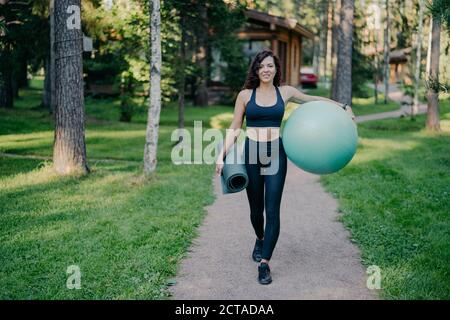 Horizontaler Schuss der Sportlerin trägt Cropped Top und Leggings, trägt Fitness-Ball und gerollt Karemat, Spaziergang auf der Straße um Bäume und grünes Gras, Stockfoto