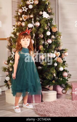 Kleines Mädchen in einem dunkelgrünen Festkleid vor dem Hintergrund eines weihnachtsbaums. Stockfoto