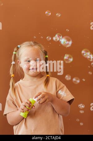 Verticalw aist up Portrait von niedlichen Mädchen mit Down-Syndrom Blasen Blasen beim Posen vor braunem Hintergrund im Studio Stockfoto