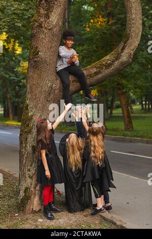 Vertikale Aufnahme der Gruppe von Kindern in halloween-Kostümen gekleidet Unter einem großen Baum stehend mit afroamerikanischem Jungen, der Süßigkeiten wirft Stockfoto