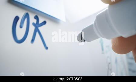 Makroaufnahme eines blauen Markierstifts, der mit der Hand gehalten wird. Person schrieb „OK“ auf einem Whiteboard mit Grafiken und Geschäftsplänen. Stockfoto
