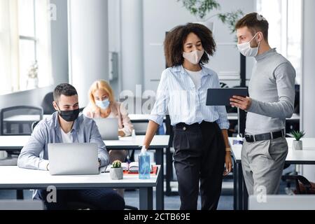 Neues Projekt und Arbeitspläne. Chef in Schutzmaske mit Tablette gibt Anweisungen für Arbeiter in Masken, afroamerikanische Frau Blick auf Gadget Stockfoto