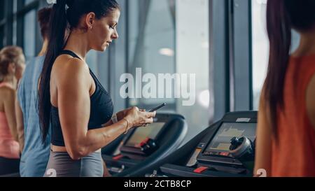 Schöne Athletic Sport Frau in der Turnhalle läuft auf einem Laufband, nutzt Smartphone. Im Hintergrund Fit Athleten Training. Seitenansicht Stockfoto