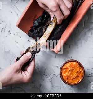 Blick aus der Perspektive eines Mannes, der eine gegrillte calcot schält, eine süsse Zwiebel, die typisch für Katalonien, Spanien ist, neben einer Steingutschüssel mit Romesco-Sauce zum Eintauchen Stockfoto