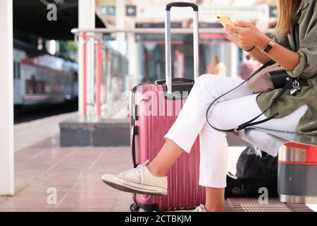 Unerkennbare junge Frau, die ihr Smartphone benutzt, während sie an einem Bahnhof auf den Zug wartet. Reisekonzept. Stockfoto