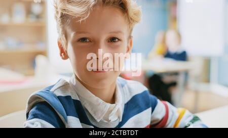 Porträt eines niedlichen kleinen Jungen mit welligen blonden Haaren sitzt an seinem Schreibtisch, smiles glücklich. Smart Little Boy mit Charming Smile sitzt in der Stockfoto