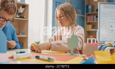 Grundschule Kunst Handwerk Klasse: Niedliche kleine Blonde Mädchen hat Spaß mit ihren Klassenkameraden auf dem Handwerk-Projekt, verwendet buntes Papier zu falten Stockfoto