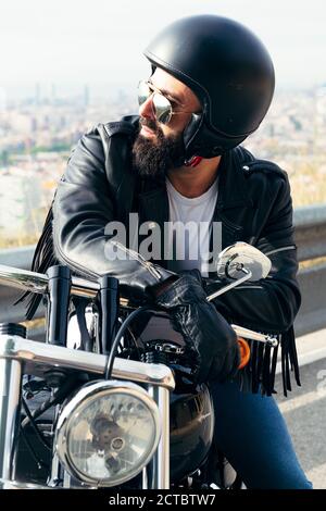 Biker mit Helm und Lederjacke sitzt auf seinem Motorrad mit der weit entfernten Stadt im Hintergrund, Freiheitsbegriff und Rebellenlebensstil Stockfoto