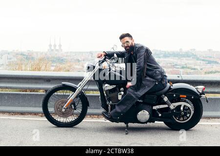 Mann in Leder und Sonnenbrille sitzend auf seinem Motorrad auf der Straße mit Panoramablick auf die Stadt, Konzept der Freiheit und Biker-Lifestyle, Stockfoto