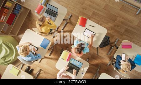 Draufsicht Aufnahme in der Grundschule Computer Science Classroom: Kinder sitzen an ihrem Schreibtisch mit Personal-Computern und digitalen Tablets für Stockfoto