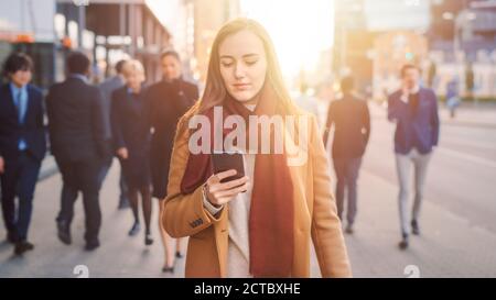 Junge lässig gekleidete Frau ist mit einem Smartphone auf einer Straße. Geschäftsleute und Büroleiter gehen Pass auf ihrem Weg zur Arbeit. Sie Sieht Aus Stockfoto