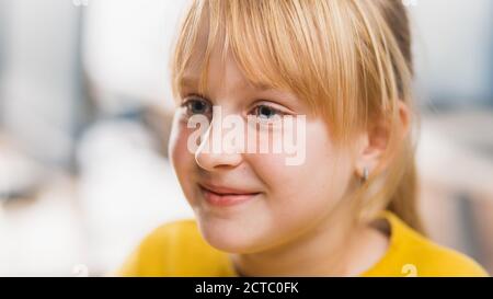 Portrait eines niedlichen kleinen Mädchens mit blondem Haar, das an ihrem Schultisch sitzt, smiles happily. Smart kleines Mädchen mit charmanten Lächeln sitzt in der Stockfoto