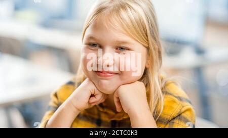 Portrait eines niedlichen kleinen Mädchens mit blondem Haar, das an ihrem Schultisch sitzt, smiles happily. Smart kleines Mädchen mit charmanten Lächeln sitzt in der Stockfoto