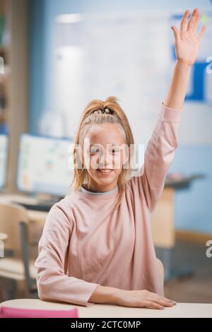 Grundschule Klassenzimmer: Porträt eines niedlichen kleinen Mädchen mit blondem Haar und Pferdeschwanz heben Hand mit einer Antwort. Brillanter junger Student fragt oder Stockfoto