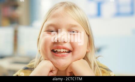 Portrait eines niedlichen kleinen Mädchens mit blondem Haar an ihrem Schultisch sitzend, Smiles happily, zeigt Zunge spöttisch. Intelligentes kleines Mädchen mit Lächeln Stockfoto