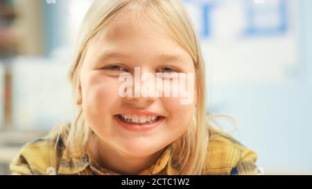 Portrait eines niedlichen kleinen Mädchens mit blondem Haar, das an ihrem Schultisch sitzt, smiles happily. Smart kleines Mädchen mit charmanten Lächeln sitzt in der Stockfoto