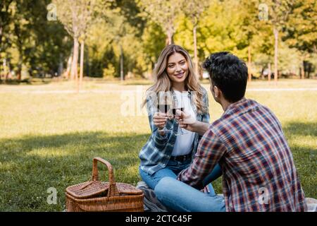 Mann und Frau sitzen auf der Decke und halten Gläser Rotwein beim Klirren im Park Stockfoto
