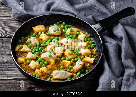 Heißer weißer Fischcurry mit grünen Erbsen und Spinat in einer Pfanne auf einem dunklen rustikalen Holztisch mit grauem Tuch, Landschaftsansicht von oben Stockfoto