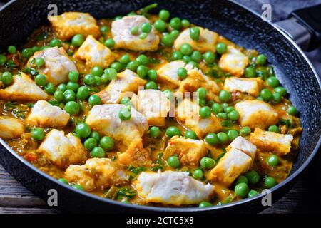 Heißer Fischcurry mit grünen Erbsen und Spinat in einer Pfanne auf einem dunklen rustikalen Holztisch mit grauem Tuch, Landschaftsansicht von oben, Nahaufnahme Stockfoto