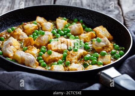 Heißer Seefischcurry mit grünen Erbsen und Spinat in einer Pfanne auf einem dunklen rustikalen Holztisch mit grauem Tuch, Landschaftsansicht von oben, Stockfoto