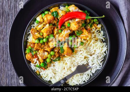 Fischcurry mit grünen Erbsen und Spinat serviert mit Reis in einer schwarzen Schüssel auf einem Holztisch, Landschaftsansicht von oben, flach liegend, Nahaufnahme Stockfoto