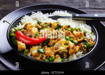 Fischcurry mit grünen Erbsen und Spinat serviert mit Reis in einer schwarzen Schüssel auf einem Holztisch, Landschaftsansicht von oben, Nahaufnahme Stockfoto