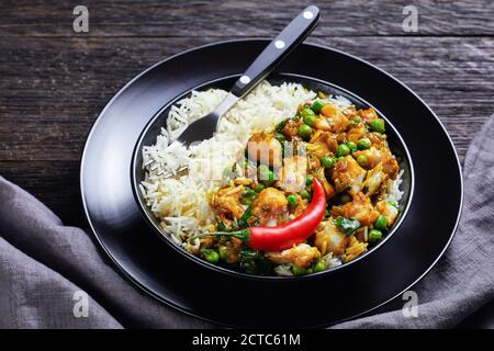 Fischcurry mit grünen Erbsen und Spinat serviert mit Reis in einer schwarzen Schüssel auf einem Holztisch, Landschaftsansicht von oben Stockfoto