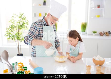 Foto von kleinen Mädchen Enkelin beobachten interessiert im Alter Opa mischen Teig in Platte fügen Sie Milch backen Kuchen Kekse zusammen alt Kochkurs am Wochenende Stockfoto