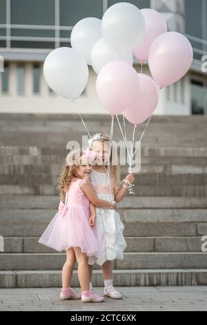 Zwei kleine Mädchen in rosa und weißen Kleidern mit Luftballons Umarmen auf der Straße Stockfoto