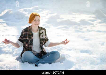 Schöne junge Frau meditiert im Winterpark. Niedlichen Ingwer Mädchen im Lotus Pose im Freien. Ruhe- und Konzentrationskonzept mit Kopierraum. Stockfoto