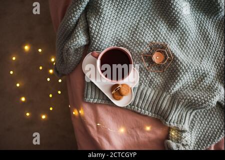 Tasse frischen Kaffee mit leckeren Keksen mit Kerze und glühenden Lichtern auf Strickpullover im Bett aus nächster Nähe. Guten Morgen. Frühstückszeit. Herbstsaison. Stockfoto