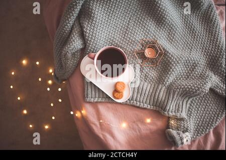 Tasse frischen schwarzen Tee oder Kaffee mit leckeren Keksen mit Kerze und glühenden Lichtern auf gestricktem Wollpullover im Bett aus nächster Nähe. Guten Morgen. Frühstück ti Stockfoto