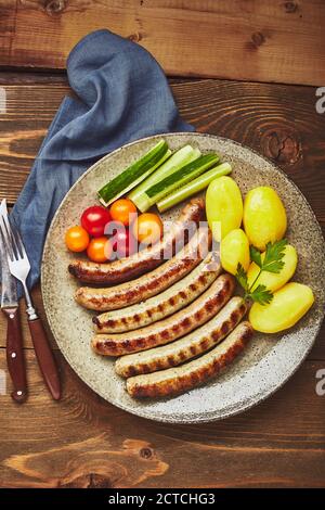 Asty gegrillte Würstchen mit Kartoffeln und Gemüse auf dem Teller in einer Nahaufnahme auf Holz Hintergrund. Draufsicht, flach liegend Stockfoto