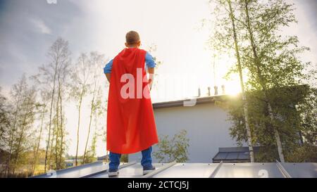 Boy spielt die Rolle eines Superhelden. Er steht auf einem Dach eines Hauses mit seinen Händen auf seiner Taille. Der junge Mann trägt ein leuchtendes rotes Cape. Er ist es Stockfoto