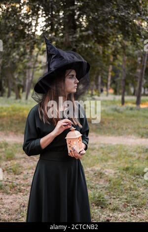 Junge Frau in dunklem Kleid und Hexenhut hält Laterne mit Kerzen in den Händen und beleuchtet den Wald. Halloween Party Kostüm. Parken mit Autum Stockfoto