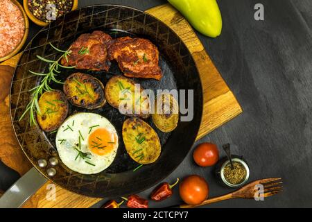 Nahaufnahme von gebratenen Schweinekoteletts in der Pfanne auf dem mit Rosmarin geschmückten Herd. Schweinehals mit Ei und Kartoffeln. Gebratenes Fleisch. Stockfoto