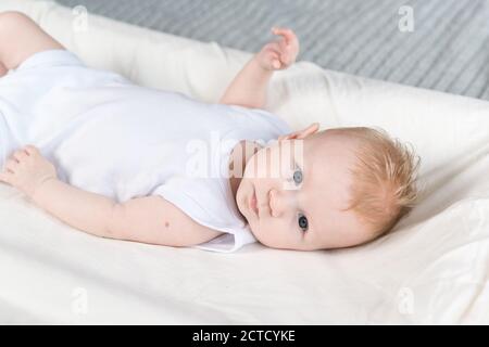 Ein vier Monate altes Baby in weißer Kleidung liegt auf dem Rücken auf einem Wickeltisch und schaut weg. Das Baby ist gut gelaunt Stockfoto