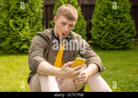 Verblüfft und erstaunt junger Mann Blick auf Smartphone sitzen auf Grünes Gras Stockfoto