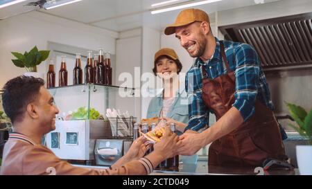 Mitarbeiter von Food Truck gibt Burger und kaltes Getränk an einen glücklichen jungen Hipster-Kunden weiter. Commercial Truck verkauft Street Food in moderner cooler Nachbarschaft Stockfoto