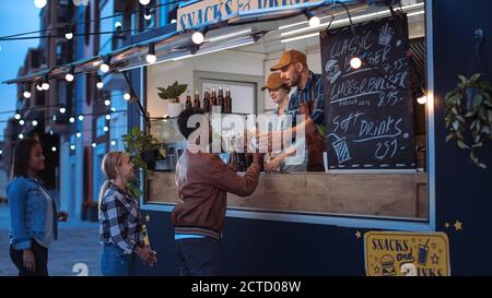 Mitarbeiter von Food Truck übergibt einem jungen Mann in Lederjacke eine Limonade und einen Burger. Der indische Mann verwendet die kontaktlose Bankkreditkarte, um für Nahrung zu zahlen Stockfoto