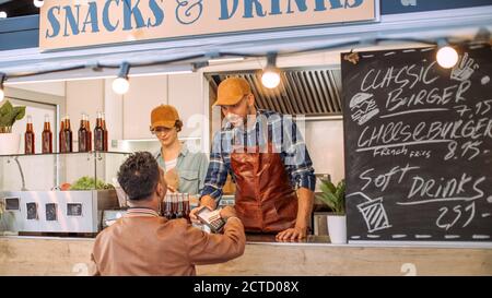 Junger indischer Mann in Lederjacke verwendet kontaktlose Bankkreditkarte, um für Nahrung zu zahlen. Street Food Truck Verkauf Burger Im Freien. Stockfoto