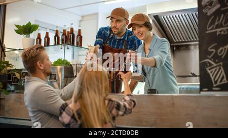 Food Truck Mitarbeiter gibt frisch zubereitete Rinderburgers, Pommes und kalte Getränke an glückliche junge Hipster-Kunden. Commercial Truck Verkauf Street Food Stockfoto